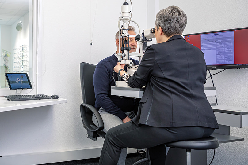 Eine Augenoptikerin führt mit einem Kunden ein Screening des vorderen Augenabschnitts durch, unterstützt durch eine Mikroskopie-Spaltlampe. Diese Untersuchung ermöglicht eine gründliche Beurteilung der Augengesundheit.